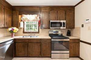 A kitchen with dark wood cabinets, a stainless steel stove and microwave, a double sink under a window with a floral valance, a bouquet of flowers on the counter, and a knife block next to the stove.