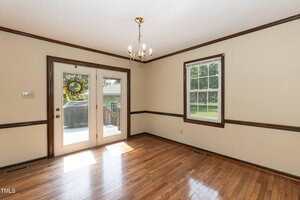 Empty room with light-colored walls, hardwood floors, and dark wood trim. A window and glass double doors provide natural light; a small gold chandelier hangs from the ceiling. Deck and greenery visible outside.