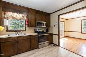 A kitchen with dark wood cabinets, beige countertops, a stainless steel stove and microwave, a double sink under a window with a floral valance, and an adjacent dining room with wood floors and a chandelier.