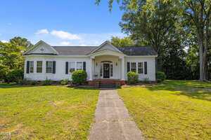 A single-story white house with black shutters at 1000 College Street, Oxford, features a covered front porch supported by columns, brick steps, and a concrete walkway winding through a green lawn with trees and shrubs.