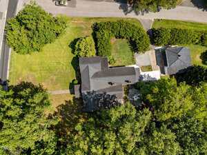 Aerial view of a house with a dark roof surrounded by trees and lawns, adjacent to a road. Another building is visible nearby, with driveways and hedges separating the properties.