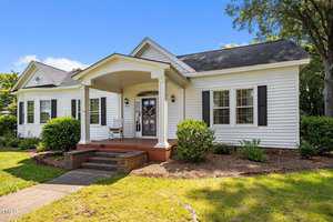 A white single-story house with black shutters, a covered front porch with columns, brick steps, and a front yard with bushes and trees under a clear blue sky.