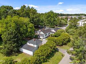 Aerial view of a single-story white house with black roof, surrounded by trees and greenery, located near a road and other buildings under a partly cloudy blue sky.