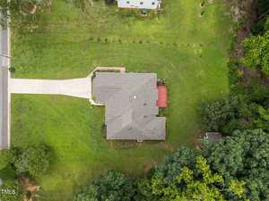 Aerial view of a single-story house with a gray roof, driveway, and red deck in a spacious, green yard. Trees border part of the property, and another structure is partially visible nearby.