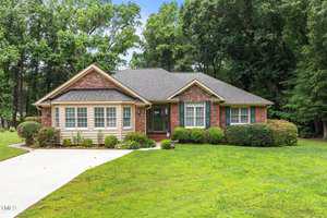 A single-story brick house with beige trim sits on a green lawn at 143 Cedarwood Drive, Henderson. Bordered by shrubs and trees, it features multiple front windows, a small porch, and a concrete driveway leading up to the entrance.