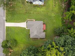 Aerial view of a single-story house with a gray roof, large green lawn, driveway leading to the street, and a red deck in the backyard. Trees and neighboring properties surround the house.