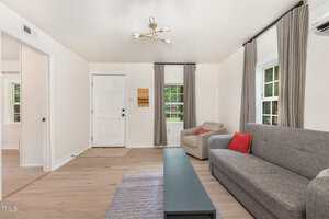 A living room with light wood flooring, a gray sofa with a red cushion, a beige armchair, a rectangular coffee table, two windows with gray curtains, and a white front door. A modern ceiling light fixture is overhead.