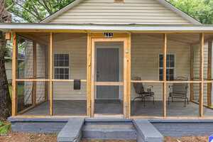 A beige house with a screened-in front porch, two metal chairs, a small table, and steps leading up to a gray front door. The porch is supported by wooden frames, and there are trees nearby.