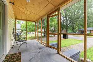 A screened-in porch with two metal chairs, a doormat, and a wooden ceiling, overlooking a grassy yard and a residential street with parked cars and trees.