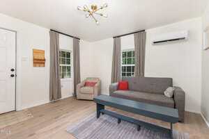 A living room with a gray sofa, blue bench-style coffee table, light armchair, two windows with gray curtains, wall-mounted air conditioner, and a modern ceiling light fixture. Neutral tones and wood flooring.