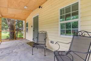 A covered porch with a concrete floor, yellow siding, a gray door, two metal chairs, a doormat, and two windows. A screened wall reveals green trees and a chain-link fence outside.