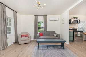 A living room with light wood flooring, a gray sofa, a beige armchair, a dark rectangular coffee table, and a rug. There are two windows with gray curtains, an air conditioner, and an open view of a modern kitchen.