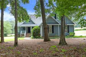 A two-story blue house with white trim and black shutters sits behind several tall trees, with a front porch, brick steps, and a landscaped yard on a sunny day.
