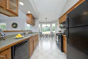 A kitchen with wood cabinets, black appliances, and a gray countertop. A bowl of lemons sits by the sink. In the background, a small dining area with two chairs and a table is positioned by large windows.