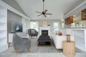 A bright living room features gray and beige furniture, a ceiling fan, a fireplace, a TV with a sailboat image, built-in shelves, and a kitchen pass-through window. The room is virtually staged.