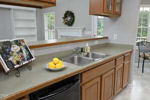 A kitchen countertop with a double sink, a bowl of lemons, a bottle of soap, and an open cookbook on a stand. Wooden cabinets are above and below the counter, and a window and wreath are visible in the background.