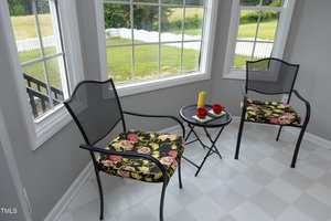 Two black metal chairs with floral cushions and a small round table with two red cups and a yellow thermos are set in a sunroom with large windows overlooking a grassy yard and white fence.