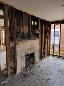 A partially demolished room with exposed wooden wall studs and a brick fireplace. Debris is scattered on the floor, and there is a view of a brick building through a window on the right.