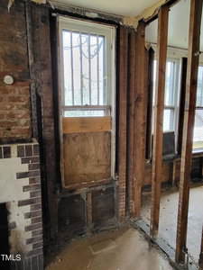 An unfinished interior wall with exposed wooden studs, a boarded-up window below a barred window, and part of a brick fireplace. Another window and open doorway are visible in the adjacent area.