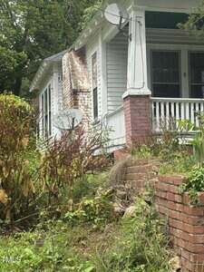 An old white house with a brick foundation and porch, a damaged and leaning brick wall on the side, overgrown plants in the yard, and two satellite dishes attached to the house.