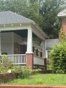 A house with a partially enclosed front porch, supported by brick and white columns, shows exposed wood and missing siding. Overgrown bushes and greenery surround the porch and brick planter in front.