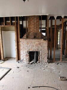 A room under renovation with exposed wall studs, a partially demolished brick fireplace in the center, debris on a wooden floor, and an open doorway to the left.