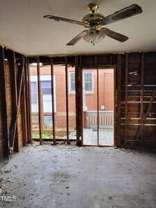 A room with exposed wall studs and no insulation or drywall, a bare concrete floor, a ceiling fan with light, and an open doorway leading to a deck outside with a view of a brick building.
