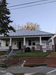 A single-story house with a gray roof, white siding, and a large front porch with white railings and brick columns. There are steps leading to the porch and a brick retaining wall in front.