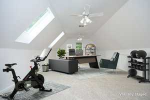 A bright attic room with sloped ceilings, a large skylight, and white walls, staged as a home office and workout area with a desk, seating, stationary bike, yoga mats, and storage racks.