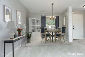 A dining room with a round table and four blue chairs on a rug, a white hutch, console table with lamps and decor, gray walls with framed art, chandelier, large window with dark curtains, and a white front door.