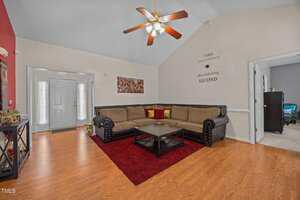 A living room with wood flooring, a brown sectional sofa, a black coffee table on a red rug, wall art, and a ceiling fan with lights. An entryway and a doorway to another room are visible.