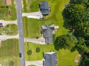 Aerial view of a suburban neighborhood showing three houses with driveways, cars parked near homes, well-maintained lawns, and trees along the right side of the image. Paved road runs vertically on the left side.