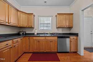 A kitchen with wooden cabinets, black countertops, stainless steel dishwasher, double sink under a window, coffee maker, roll of paper towels, and a red mat on a wood floor. Doorway visible to the right.