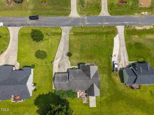 Aerial view of three houses with driveways leading to a suburban street. The yards are green and well-maintained. One white car is parked in a driveway, and a black car is driving on the road. Trees and small bushes are visible.