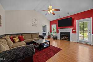 A living room with wood flooring, a sectional sofa, a coffee table, a red accent wall, a fireplace with a mounted TV above it, a ceiling fan, and glass doors leading outside.