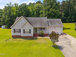 Located at 106 Oak Road in Butner, this single-story gray suburban house features white trim, a brick foundation, and an attached two-car garage. It sits on a large grassy lot with trees in the background and a driveway to the right.