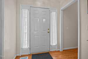A white front door with two narrow windows on each side covered by sheer white curtains, set in a beige entryway with hardwood floors and a black doormat. An open doorway leads to another room.