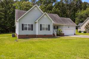 Located at 106 Oak Road in Butner, this single-story house features light gray siding, black shutters, a brick foundation, a two-car garage, a charming front porch with white railings, and a spacious green lawn bordered by trees.