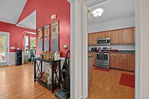A kitchen with stainless steel appliances and wooden cabinets is visible through a doorway. The adjacent room has red walls, a drink station, a vacuum cleaner, and large windows letting in natural light.