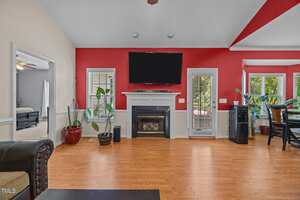 A living room with red and beige walls, a fireplace below a wall-mounted TV, two windows, potted plants, a door leading outside, and hardwood floors. A dining area and a bedroom are visible in the background.