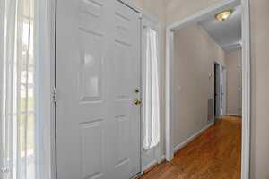 A view of a home's entryway with a closed white front door, gold door hardware, light-colored walls, and wood flooring. A hallway extends to the right, featuring white trim and a ceiling light.