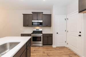 Modern kitchen with stainless steel gas stove and microwave, dark wood cabinets, white countertops, a sink on an island, light-colored walls, wooden floor, and two white paneled doors.