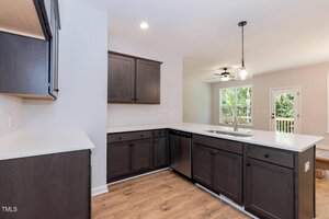 Modern kitchen with dark wood cabinets, white countertops, stainless steel appliances, light wood flooring, and an island sink. The space opens to a bright living area with large windows and a door leading outside.