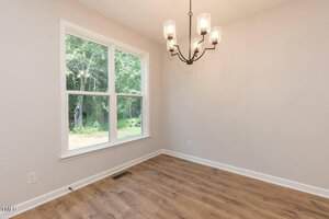 A small empty room with light-colored walls, a large window showing trees outside, wood flooring, and a black metal chandelier with five lights hanging from the ceiling.