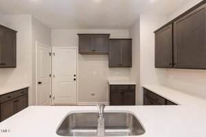 A modern kitchen with gray cabinets, white walls, white countertops, a double sink with a faucet in the foreground, and a white door in the background. There are no appliances visible.