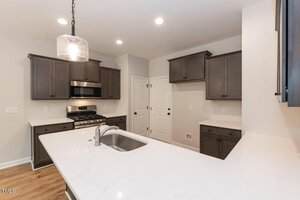 A modern kitchen with dark cabinets, white countertops, a stainless steel gas stove and microwave, a central island with a sink, wood flooring, and recessed ceiling lights.