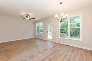 A bright, empty room with light-colored walls, wood flooring, large windows, a ceiling fan with lights, and a glass door leading outside. A black chandelier-style light fixture hangs from the ceiling.