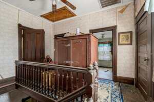 A hallway with patterned wallpaper and wooden trim features a large wardrobe, a stair railing, and several doors. The ceiling has a boarded-up opening, and the adjacent room shows peeling paint and a blue patterned rug.