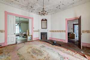 A worn, unfurnished room with a floral rug, peeling ceiling paint, a fireplace with a mirror, a chandelier, and two open doorways, revealing adjoining rooms with vintage furniture and house items.