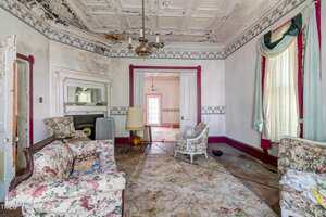 A worn living room with old floral sofas, a fireplace, a standing lamp, peeling paint on the ceiling, stained walls, and a large window with curtains. The floor is bare, and another room is visible through a doorway.